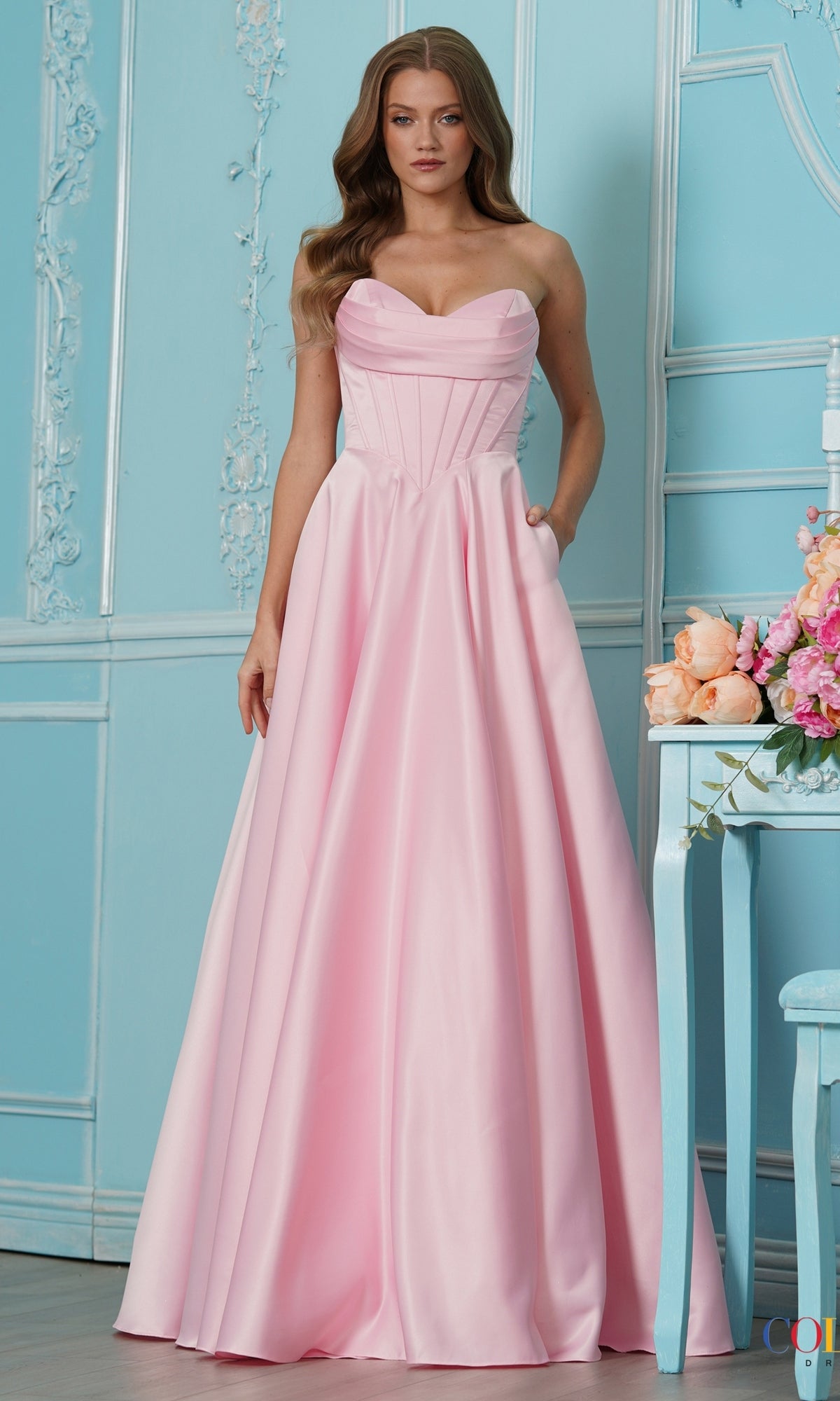 A model wearing the pink Long Prom Dress: Colors Dress 4063, a strapless, floor-length satin A-line gown with a pleated sweetheart neckline, corset-style bodice, and side pockets.